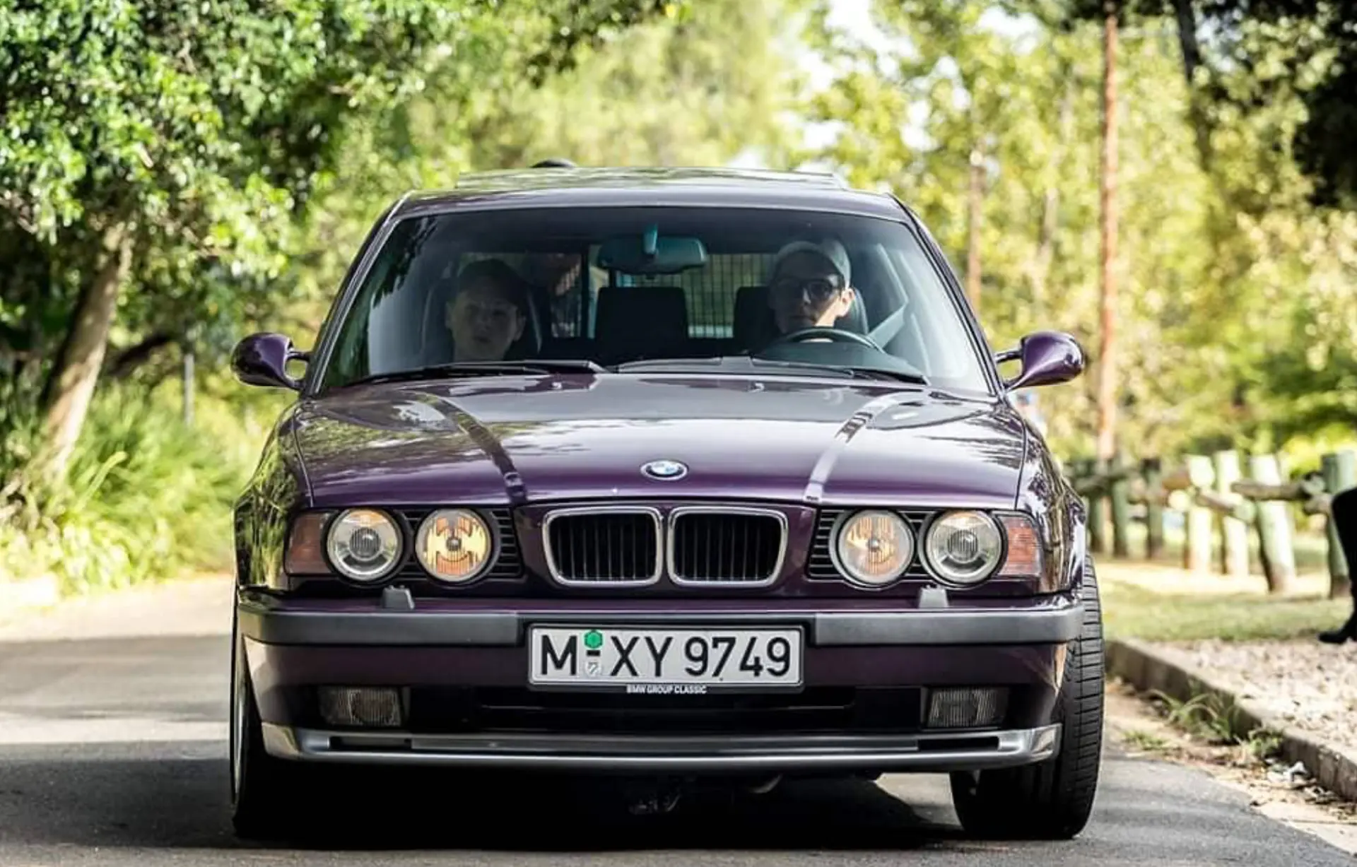 Purple BMW E34 M5 photographed head-on while driving along a tree-lined road