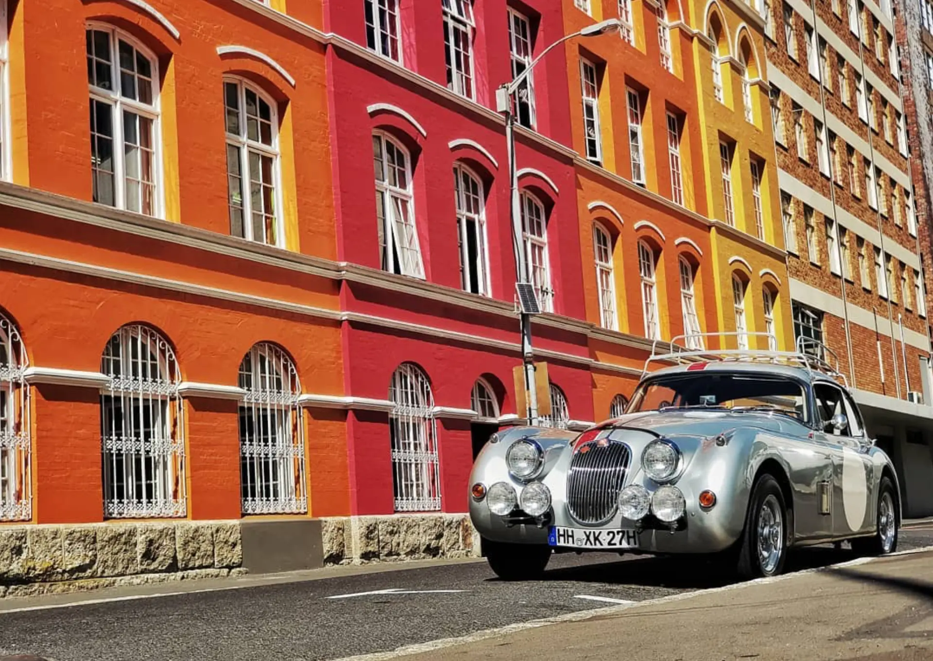 Silver classic Jaguar with rally lights driving past colourful Cape Town buildings