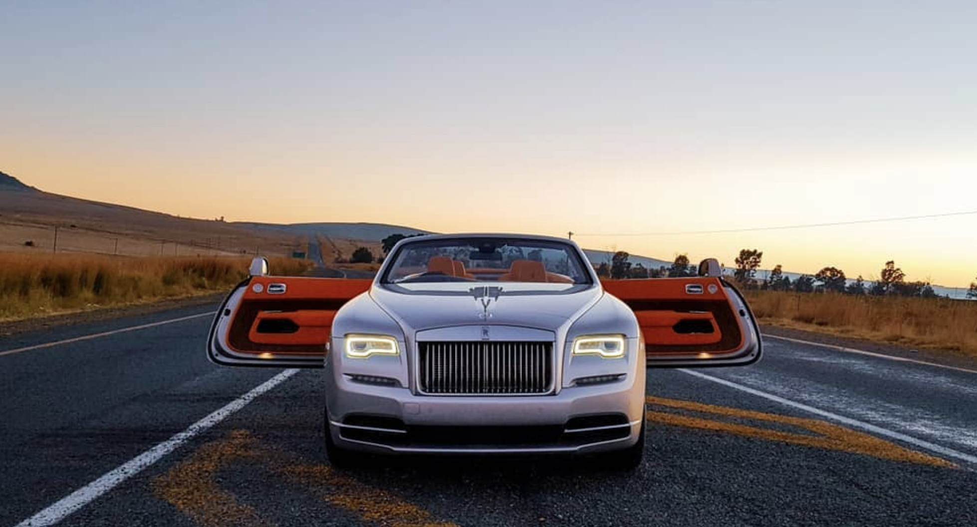 Silver Rolls-Royce Dawn facing the camera on an open road at sunset with both doors open