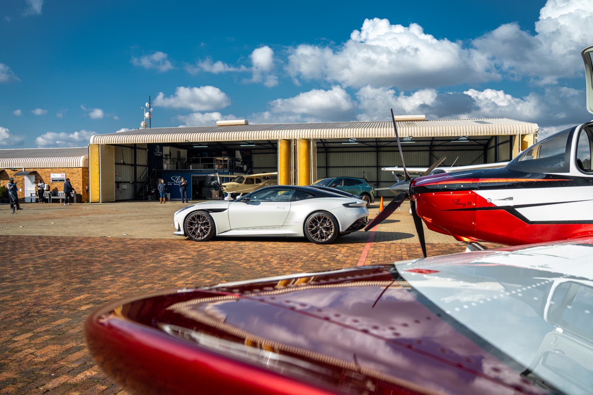 White Aston Martin parked among light aircraft outside an airport hangar