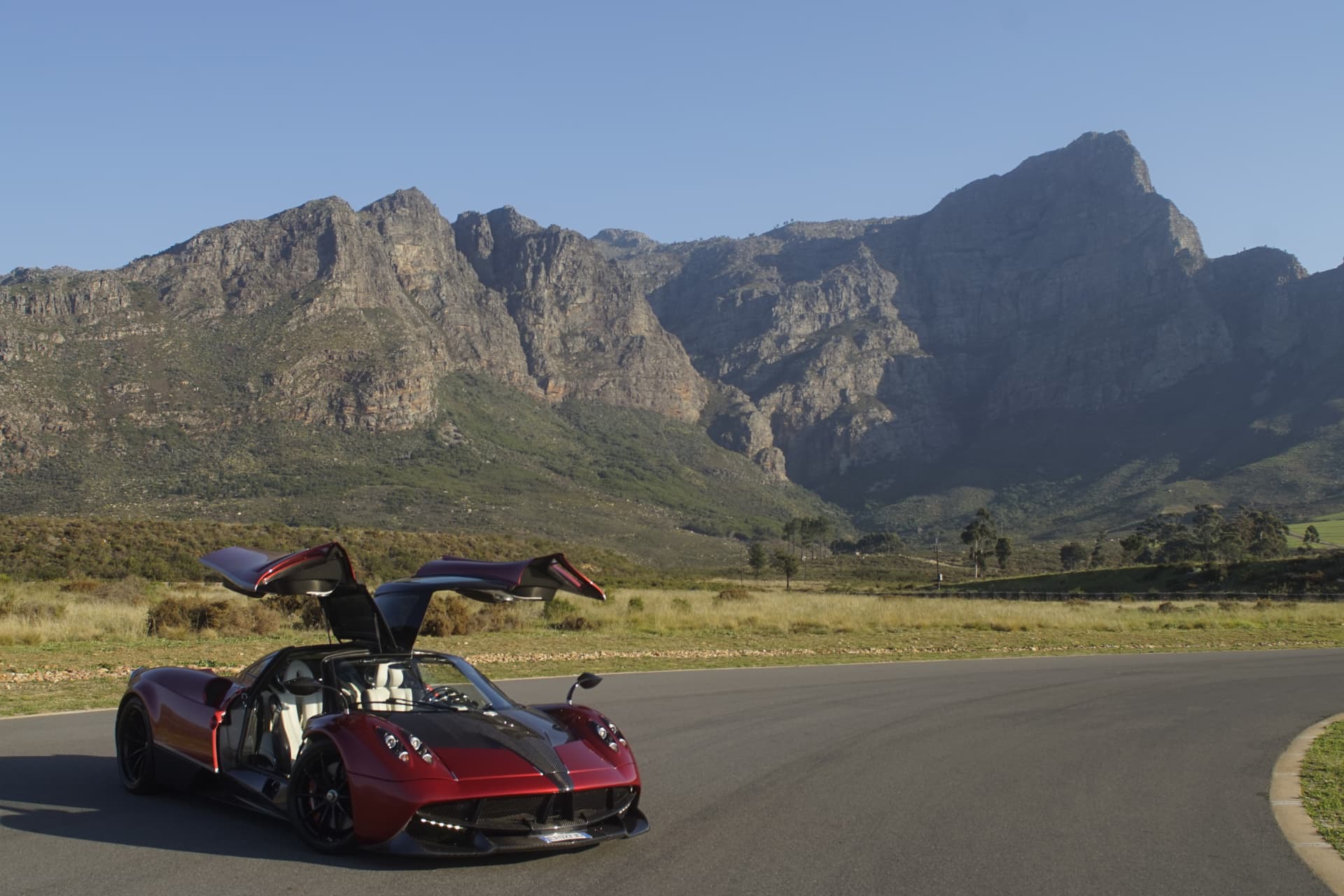 Red Pagani Huayra prototype parked on a mountain pass with both gullwing doors raised, rocky peaks behind