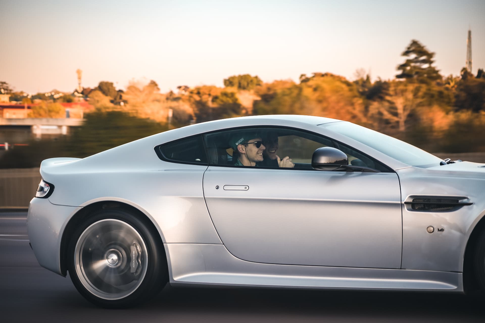 Silver V12 Vantage at speed in evening light with two smiling occupants on board