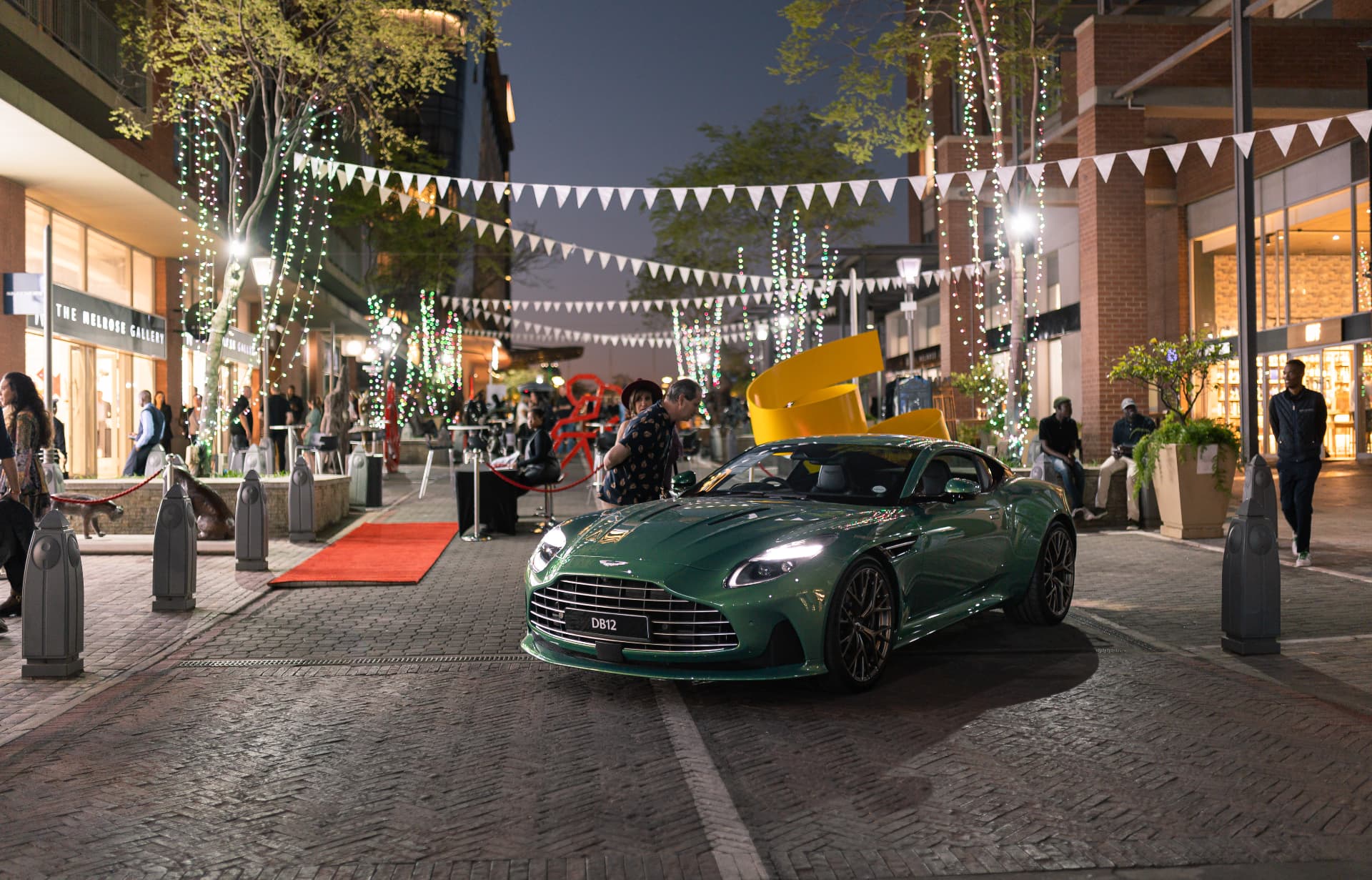 Green DB12 parked on the Melrose Arch High Street at night among bunting and sculptures